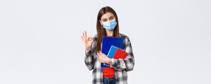 Coronavirus pandemic, covid-19 education, and back to school concept. Determined young woman, student in medical mask with notebooks and study material, show okay, no problem sign.