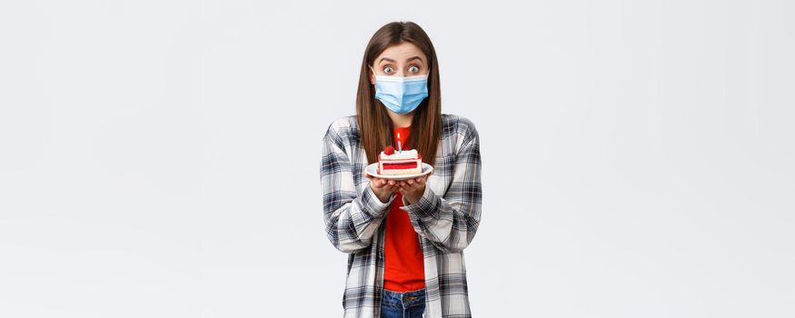 Coronavirus outbreak, lifestyle during social distancing and holidays celebration concept. Cute happy birthday girl making wish, wear medical mask, holding b-day cake, celebrating inside home.