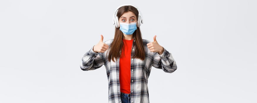 Social distancing, leisure and lifestyle on covid-19 outbreak, coronavirus concept. Enthusiastic upbeat girl in headphones and medical mask, show thumb-up like awesome music sound, white background.