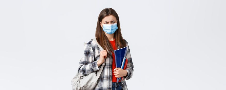 Coronavirus pandemic, covid-19 education, and back to school concept. Upset disappointed female student in medical mask hate studying in university during infection oubreak, frowning condemn dean.