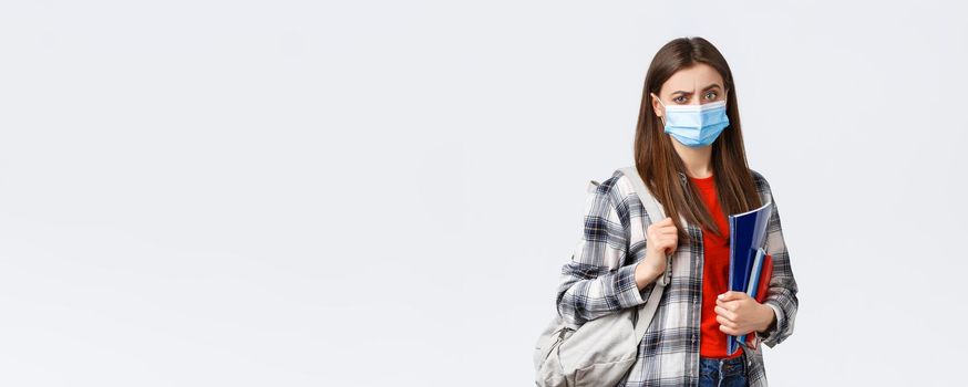 Coronavirus pandemic, covid-19 education, and back to school concept. Doubtful and skeptical female student in medical mask frowning disappointed, hold notebooks and backpack.