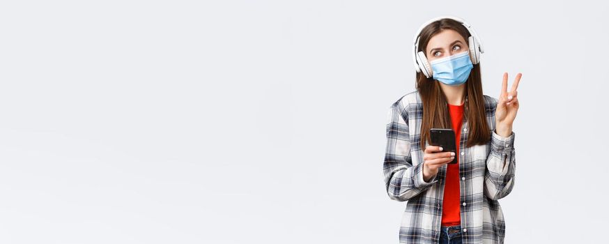 Social distancing, leisure and lifestyle on covid-19 outbreak, coronavirus concept. Happy cheerful silly girl in medical mask and headphones, show peace sign smiling, holding mobile phone.