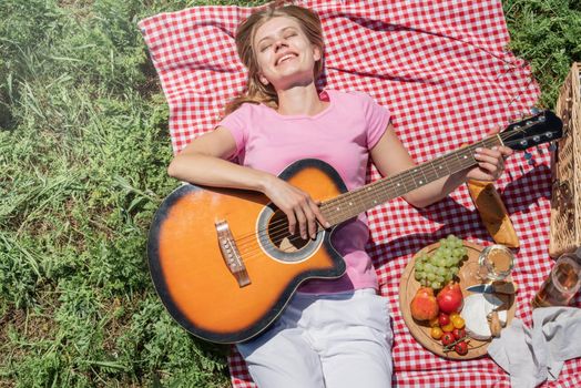 Top view of young caucasian woman in white pants outside having picnic, eating and playing guitar. Summer fun and leisure