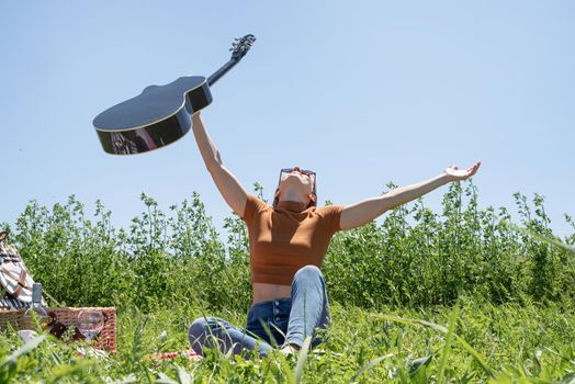 millennial woman in summer clothes and sunglasses playing guitar on a picnic. Happy woman raising her hands to the sky, holding guitar