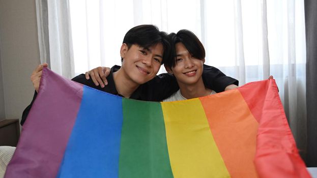 Cheerful gay couple embracing and holding LGBT pride flag with love together. LGBT, pride, relationships and equality concept.