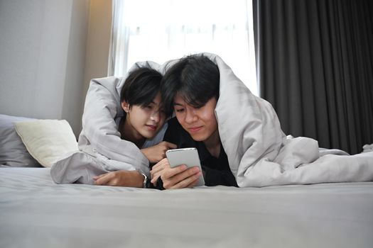 Young gay couple lying under blanket on bed and using smart phone together. LGBT and love concept.