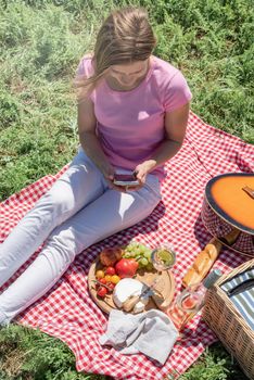 woman in white pants and pink shirt outside having picnic and using smartphone taking photo. Summer fun and leisure