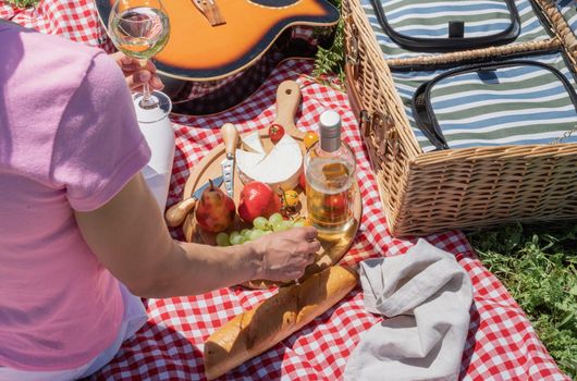 Top view of unrecognizable young woman in white pants outside having picnic, eating and playing guitar. Summer fun and leisure. view from behind