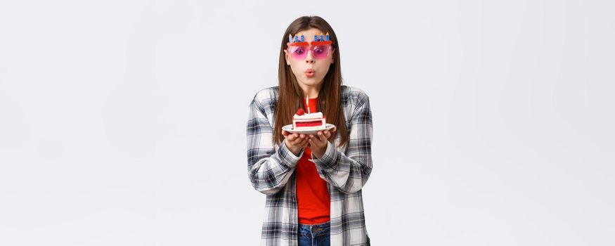 People lifestyle, holidays and celebration, emotions concept. Cute tender young b-day girl in glasses, blowing lit candle on birthday cake, making wish and celebrating at party, white background.