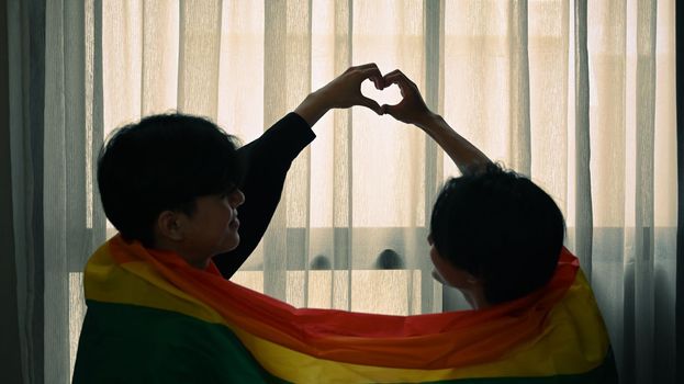 Romantic young gay couple making heart with their hands while sitting under rainbow flag. LGBT, pride, relationships and equality concept.