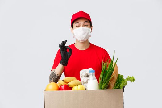 Online shopping, food delivery and coronavirus pandemic concept. Smiling asian courier in uniform show okay sign, give client his order, fresh groceries from online shop, wear medical mask.