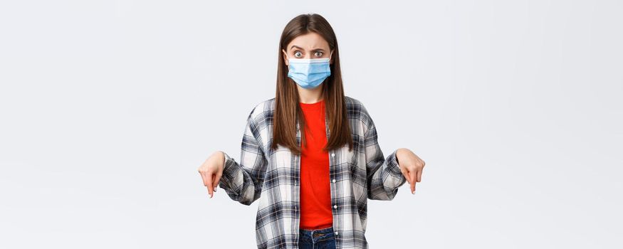 Coronavirus outbreak, leisure on quarantine, social distancing and emotions concept. Confused woman in medical mask cant understand what happening, pointing fingers down, look alarmed.