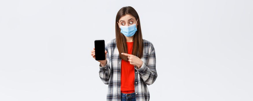 Different emotions, covid-19, social distancing and technology concept. Excited and enthusiastic good-looking girl in medical mask, pointing looking at mobile phone display, promote app.