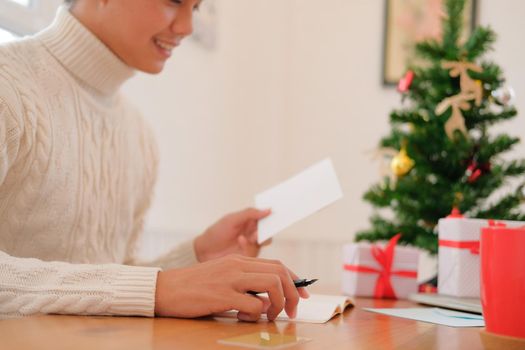 man wearing cream sweater writing christmas letter greeting card holiday wishes with xmas decoration.