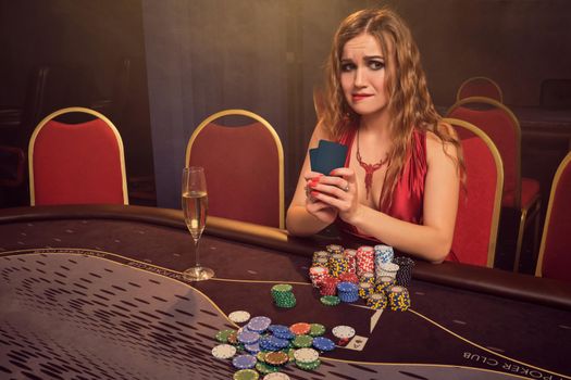 Beautiful maiden with a long curly hair and perfect make-up, dressed in a sexy red dress. She is sitting at a gambling table with two playing cards in her hands and looking unhappy. Poker concept on a dark smoke background in a ray of a yellow spotlight. Casino.