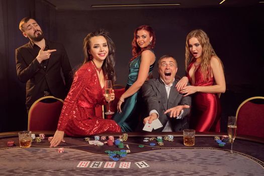 Joyful colleagues are playing poker at casino. They are celebrating their win, smiling and looking vey excited while posing at the table against a dark background. Cards, chips, money, alcohol, gambling, entertainment concept.