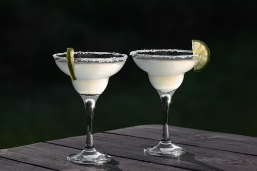Two glasses with fresh margarita cocktail on a wooden table on a patio summer evening, selective focus.