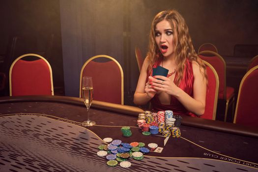 Beautiful lady with a long curly hair and perfect make-up, dressed in a sexy red dress. She is sitting at a gambling table with two playing cards in her hands and looking shocked. Poker concept on a dark smoke background in a ray of a yellow spotlight. Casino.