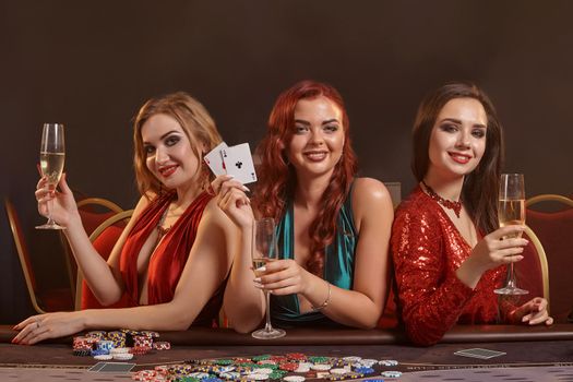 Three charming ladies in a sexy dresses are playing poker at casino. They are celebrating their win, looking at the camera and smiling, holding champagne and cards in their hands while posing sitting at the table against a smoke background. Chips, money, gambling, entertainment concept.