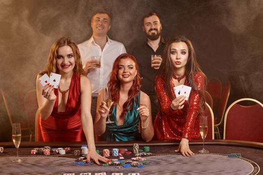 Group of a pleasant classmates are playing poker at casino. They are celebrating their win, looking at the camera and holding some playing cards in their hands while posing at the table against a smoke background. Chips, money, alcohol, gambling, entertainment concept.