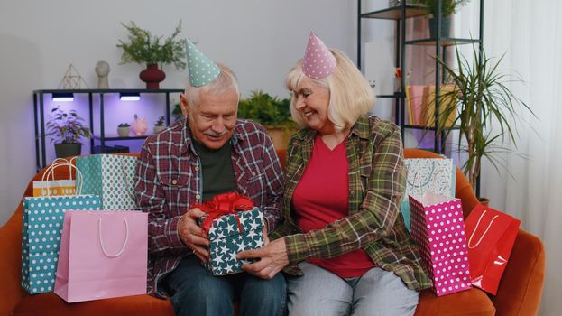 Happy old senior elderly family couple grandparents man woman celebrating birthday anniversary on couch at home. Mature grandmother wife giving gift box surprise present to her husband grandfather