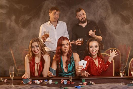 Agitated friends are playing poker at casino. They are celebrating their win, smiling and posing at the table against a dark smoke background. Cards, chips, money, alcohol, fortune, gambling, entertainment concept.