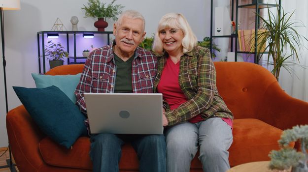 Senior elderly family couple grandparents man woman sitting on couch at home room and opens laptop. Mature grandmother grandfather starts working on computer, sends messages, makes online purchases