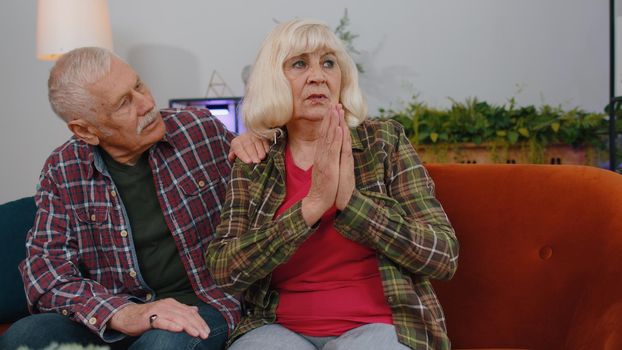 Senior elderly family couple grandparents man woman pray with hope, looking upward and making wish, asking with hopeful imploring expression, begging apology. Mature grandmother grandfather at home