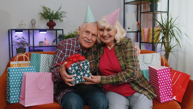 Happy old senior elderly family couple grandparents man woman celebrating birthday anniversary on couch at home. Mature grandmother wife giving gift box surprise present to her husband grandfather