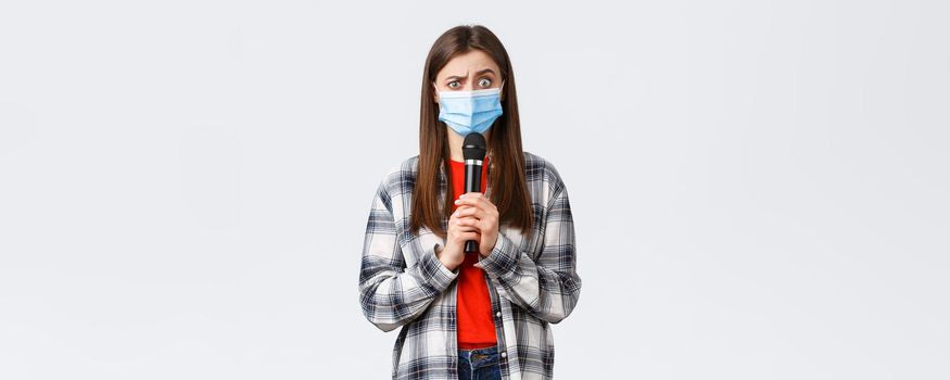 Coronavirus outbreak, leisure on quarantine, social distancing and emotions concept. Confused woman in medical mask look smth strange, holding microphone, perform, white background.