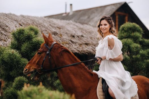 A woman in a white sundress riding a horse near a farm.