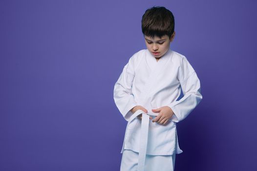 Horizontal portrait of a confident teenage boy aikido fighter tying white belt of his kimono, isolated over violet background with copy ad space. Oriental philosophy and japaneese martial arts concept