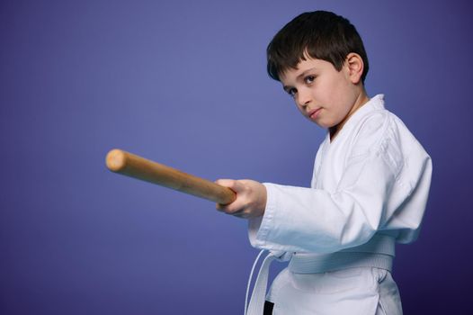 Waist length portrait of a confident teenage boy in white kimono practices wooden bokken in Aikido training on purple background with copy ad space. Concepts of Oriental martial arts practice