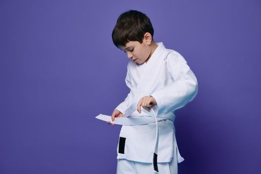 Confident Caucasian teenage boy puts on white kimono, ties a belt around his waist, ready for combat sports. Aikido fighter. Oriental martial arts. Combat sport concept