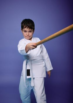 Oriental martial arts practice, combat, aikido, japanese sports and philosophy. Child boy fighter with wooden weapon bokken sword against violet background with copy space