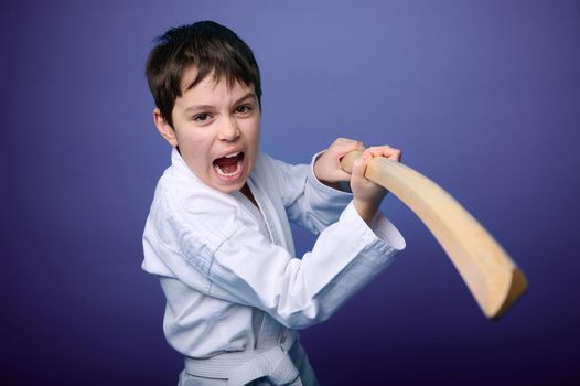 Waist length portrait of a concentrated strong Caucasian teenage boy in white kimono standing in fighting pose with wooden katana sword. Aikido fighter. Oriental martial arts concept