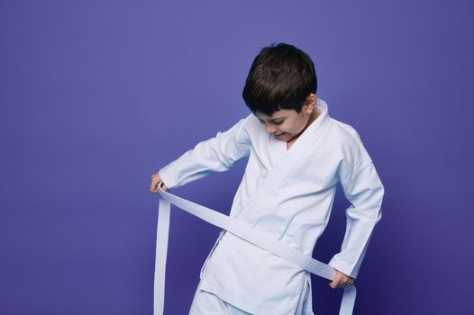 Confident and happy school age boy holding white belt of his aikido kimono and tying it around waist, isolated over purple background with copy ad space. Oriental combat sport, martial arts concept