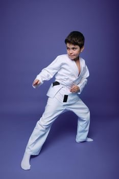 Oriental martial arts. Full length portrait of an aikido fighter, Caucasian 10 years old boy in kimono improves his fighting skills, isolated on violet background with copy space