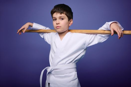 Confident charming Caucasian aikido fighter poses with wooden weapon during Oriental martial arts practice, isolated on violet background, copy ad space for text. Oriental martial arts practice