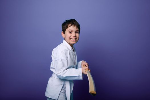 Waist length portrait of a smiling Caucasian teenage boy in white kimono practicing wooden sword in Aikido training on purple background with copy ad space. Concepts of Oriental martial arts practice
