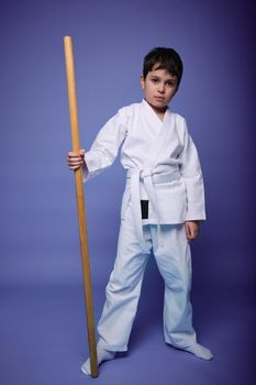 Learning aikido, Oriental martial arts concept. Caucasian Cute boy in a white kimono with a wooden weapon in his hand isolated on purple background with copy space for advertising text