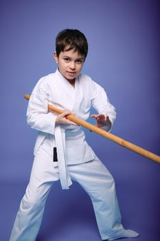 Caucasian teenage boy in a white kimono fights with a wooden sword in aikido training on a purple background with copy space for advertising text. Healthy lifestyle and martial arts concept
