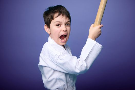 Concentrated strong boy in white samurai hakama kimono - aikido fighter holding bamboo katana weapon and practicing martial skills isolated on purple background with copy space for advertising text