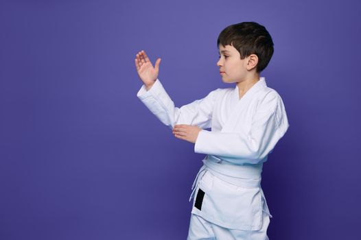 Charming confident Oriental martial arts wrestler - Caucasian school age boy in white kimono- practicing martial skills isolated on purple background with copy space for advertising text. Aikido