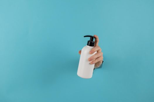 A woman's hand sticks out of a hole in a blue paper background and holds a skin lotion pump