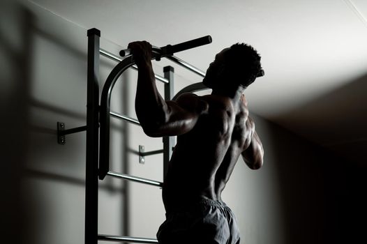 African american man with naked torso pulls up on horizontal bar in gym