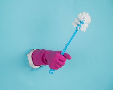 A woman's hand in a rubber glove holds a toilet brush and sticks out of a hole on a blue paper background