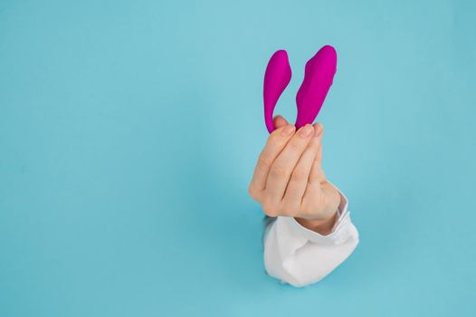 A woman's hand holds a pink sex toy through a hole in a paper blue background