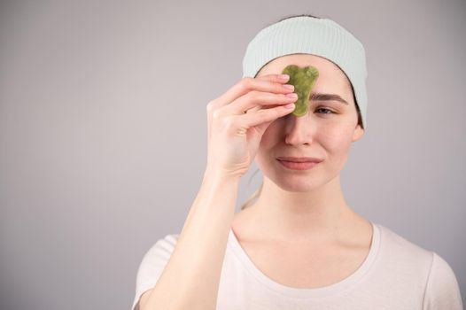 Portrait of a young woman massages her face with a gouache scraper on a white background