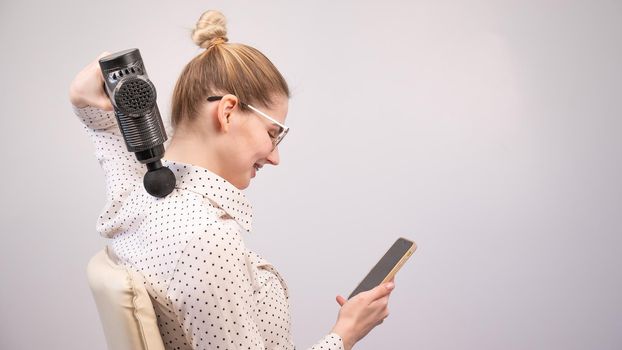 A young woman sits on a chair, uses a smartphone and makes herself massage with an electro massage gun